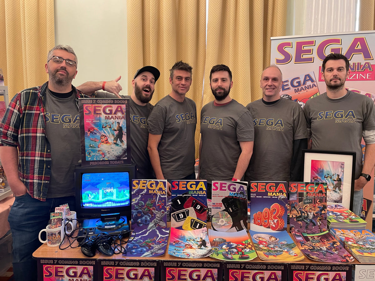 Six members of the Sega Mania team in matching Sega Mania T-shirts stand behind their table at Play Expo Blackpool, with printed issues, mugs and a Mega Drive on show.